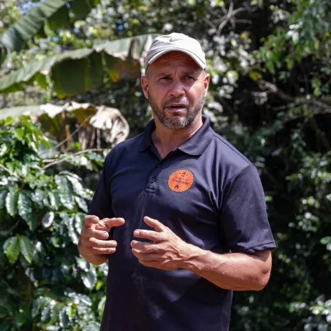 A man wearing a tan baseball cap with coffee plants and lush foliage in the background